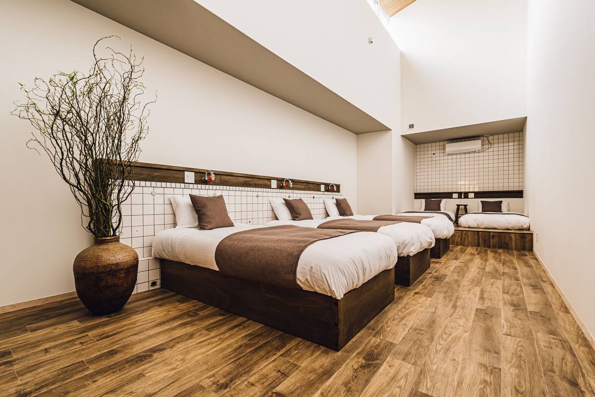 A modern hotel room with a large bed, a wooden floor, a large vase with plants, and a mirror on the wall.