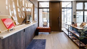 Un hall d’hôtel avec un comptoir d’accueil en bois, une brochure intitulée « Tabist », un paillasson bleu et une vue sur l’extérieur à travers de grandes fenêtres.
