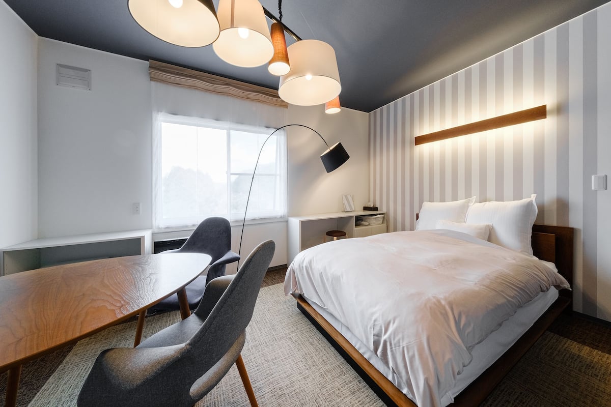 Une chambre moderne avec un grand lit, un bureau et des chaises. Une fenêtre laisse entrer la lumière naturelle, et l’une des parois est décorée de motifs rayés.