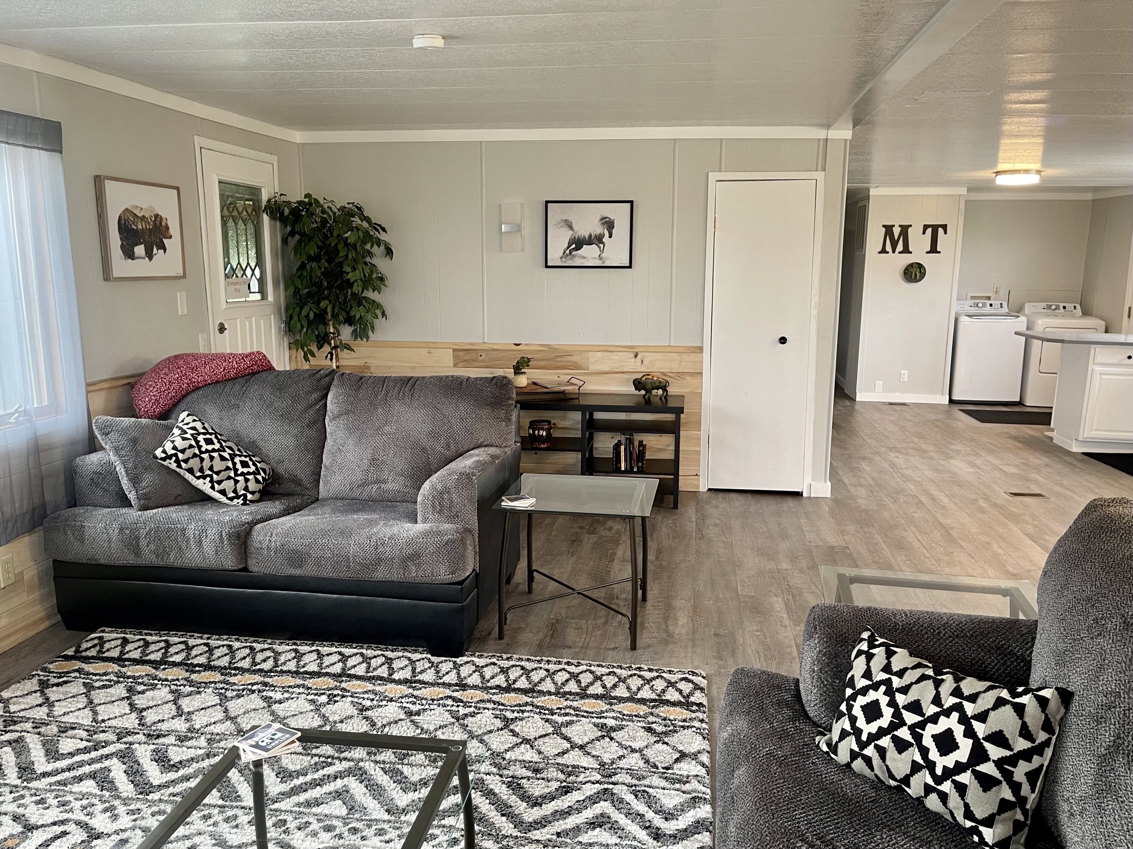 A living room with a grey sofa, a patterned rug, and a small table. There is a door leading to a kitchen area.