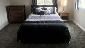 Un dormitorio con cama, mesita de noche con lámpara, cómoda y ventana con cortinas.