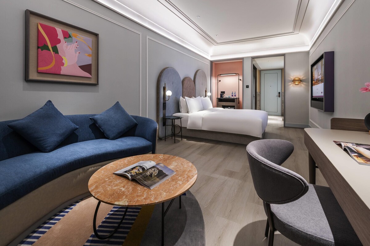A modern hotel room with a blue sofa, a round marble table, a gray armchair, a bed, and a TV mounted on the wall.