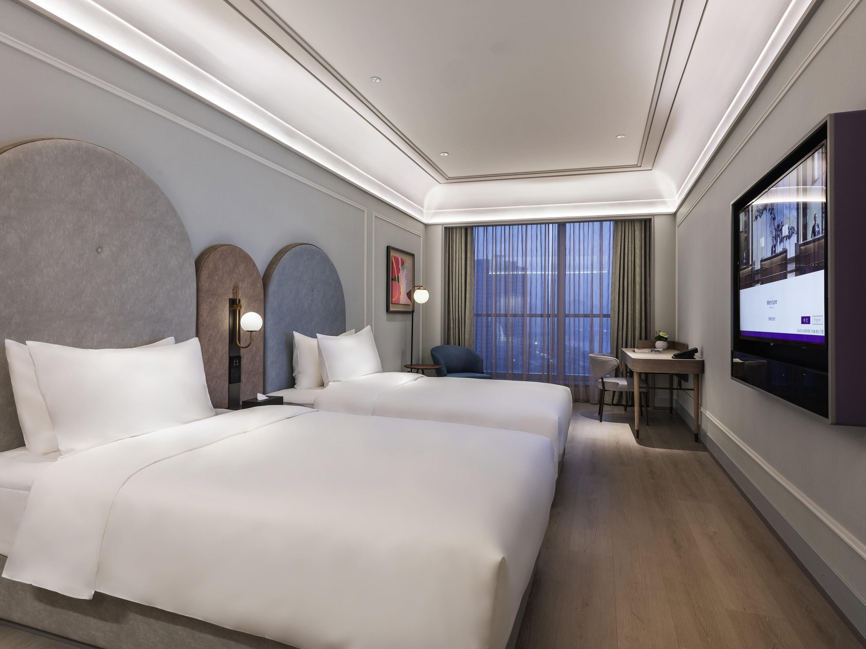 Habitación de hotel con dos camas, un escritorio, una silla, un televisor y un ventanal grande con cortinas.