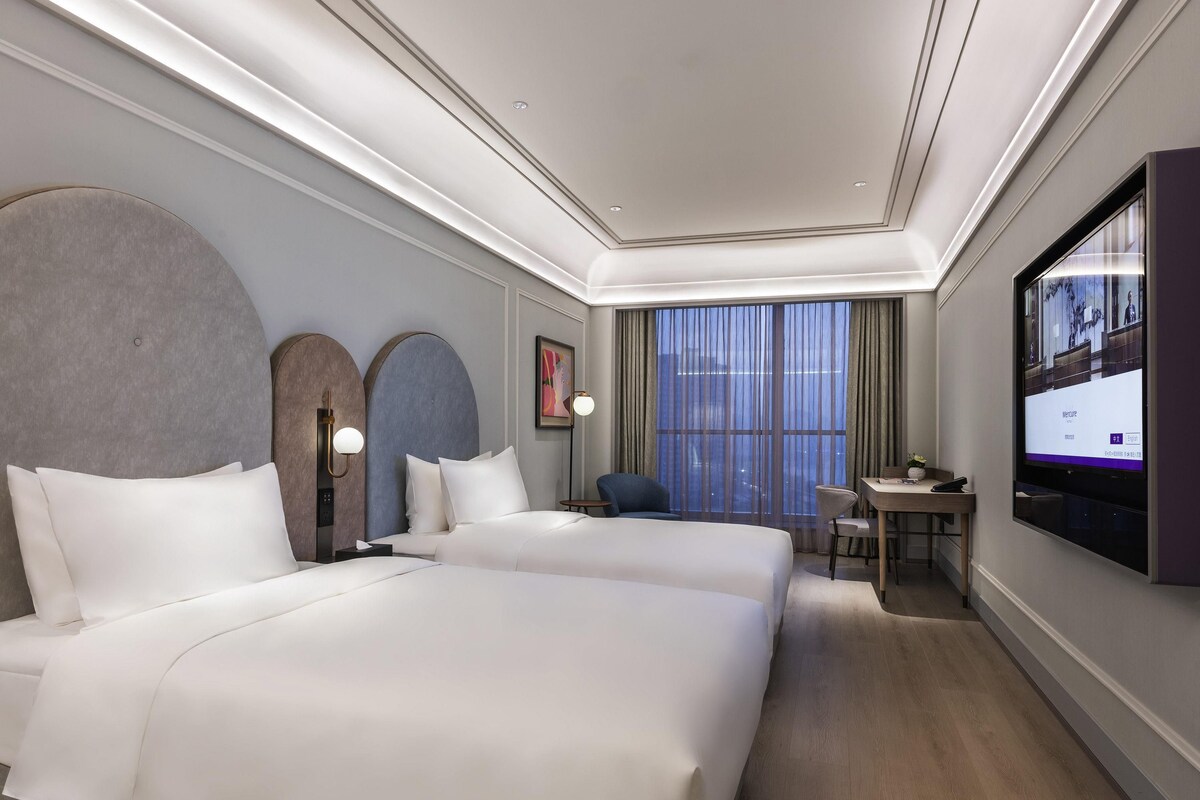 A hotel room with two beds, a desk, a chair, a TV, and a large window with curtains.