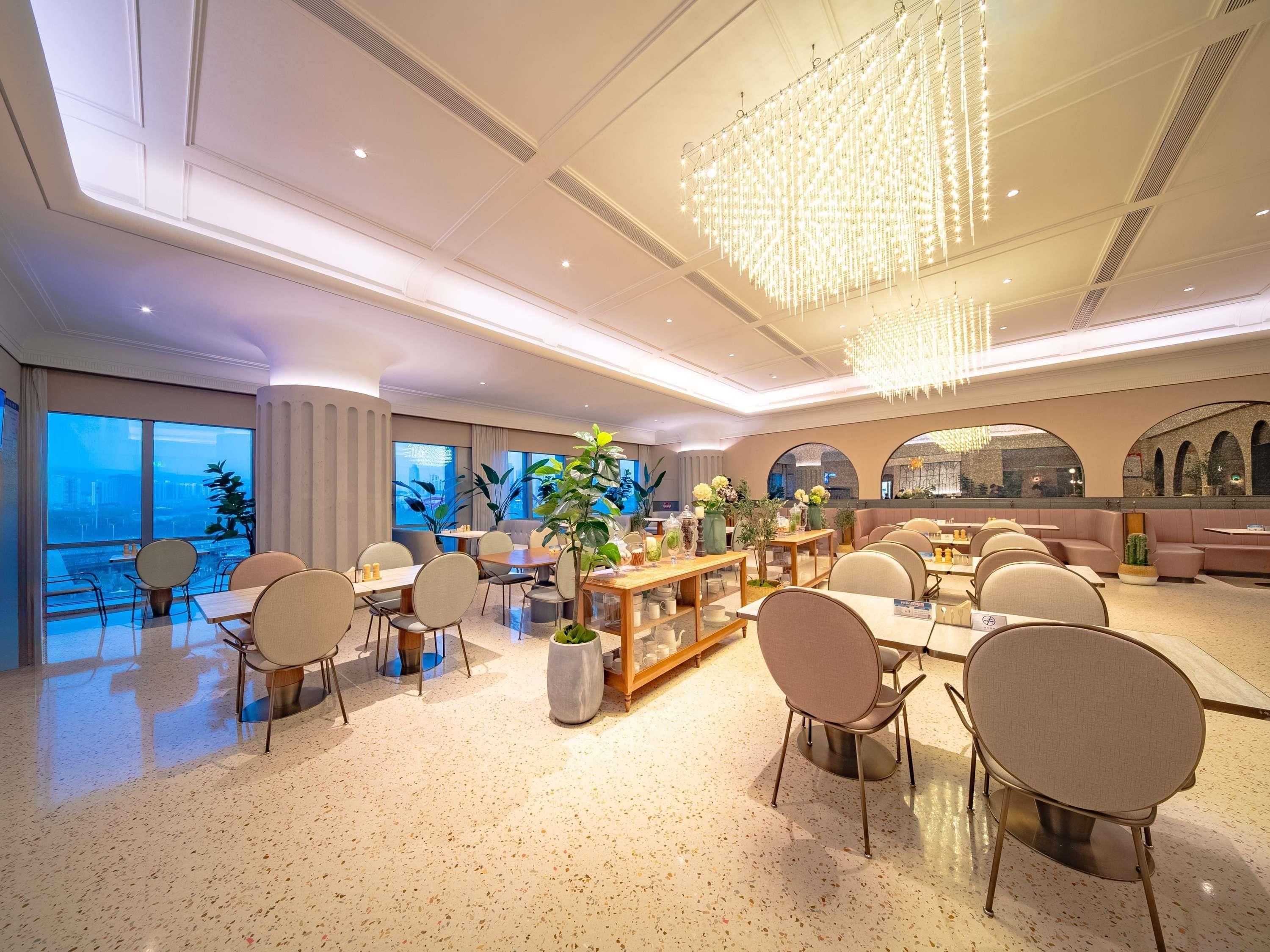 A spacious hotel dining area with multiple tables and chairs, a central bar area, and large windows offering a view of the cityscape.