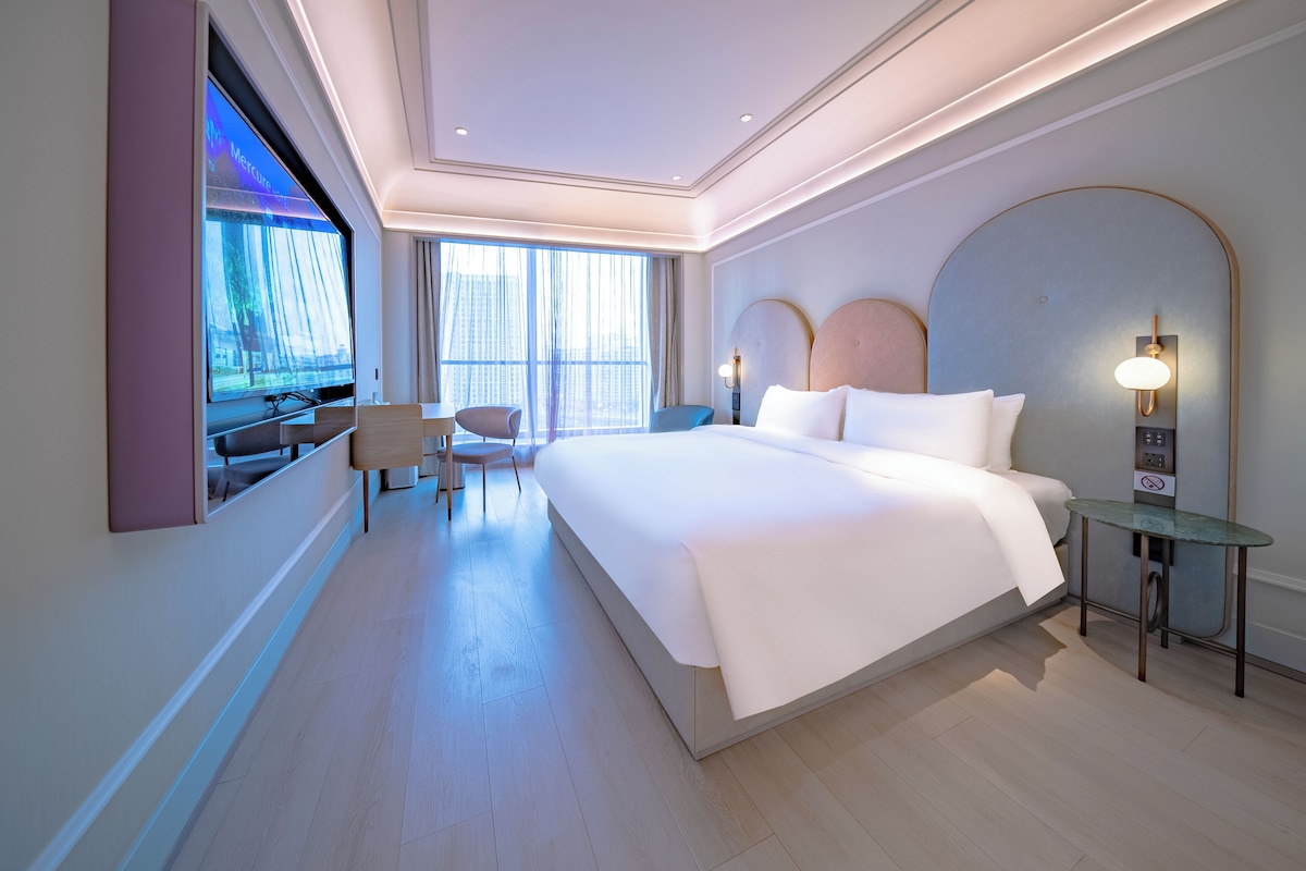 Habitación de hotel moderna con una cama grande, un escritorio, una silla, un televisor y un ventanal amplio.
