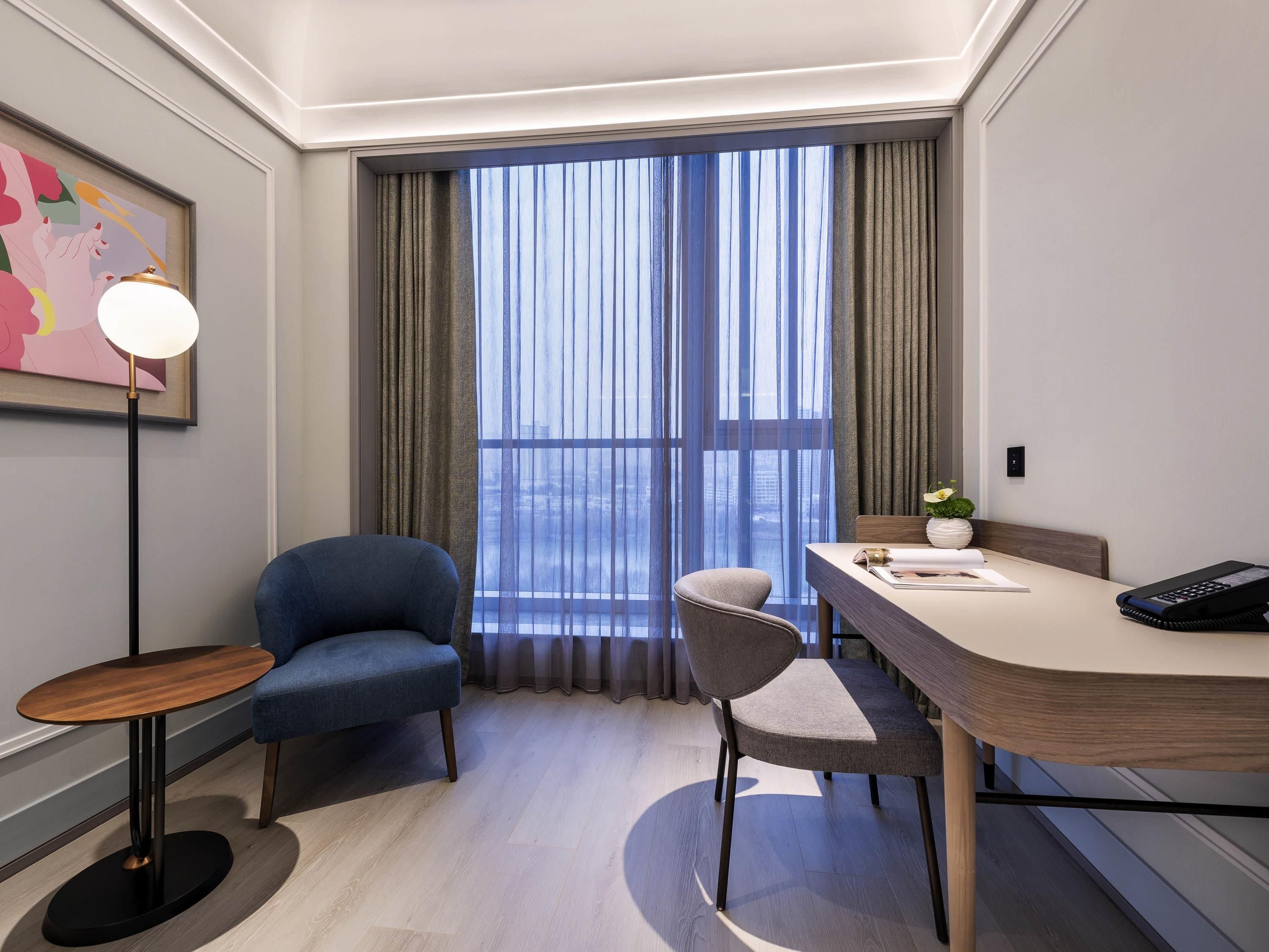A hotel room with a large window, a desk with a phone, a chair, and a small table with a lamp.