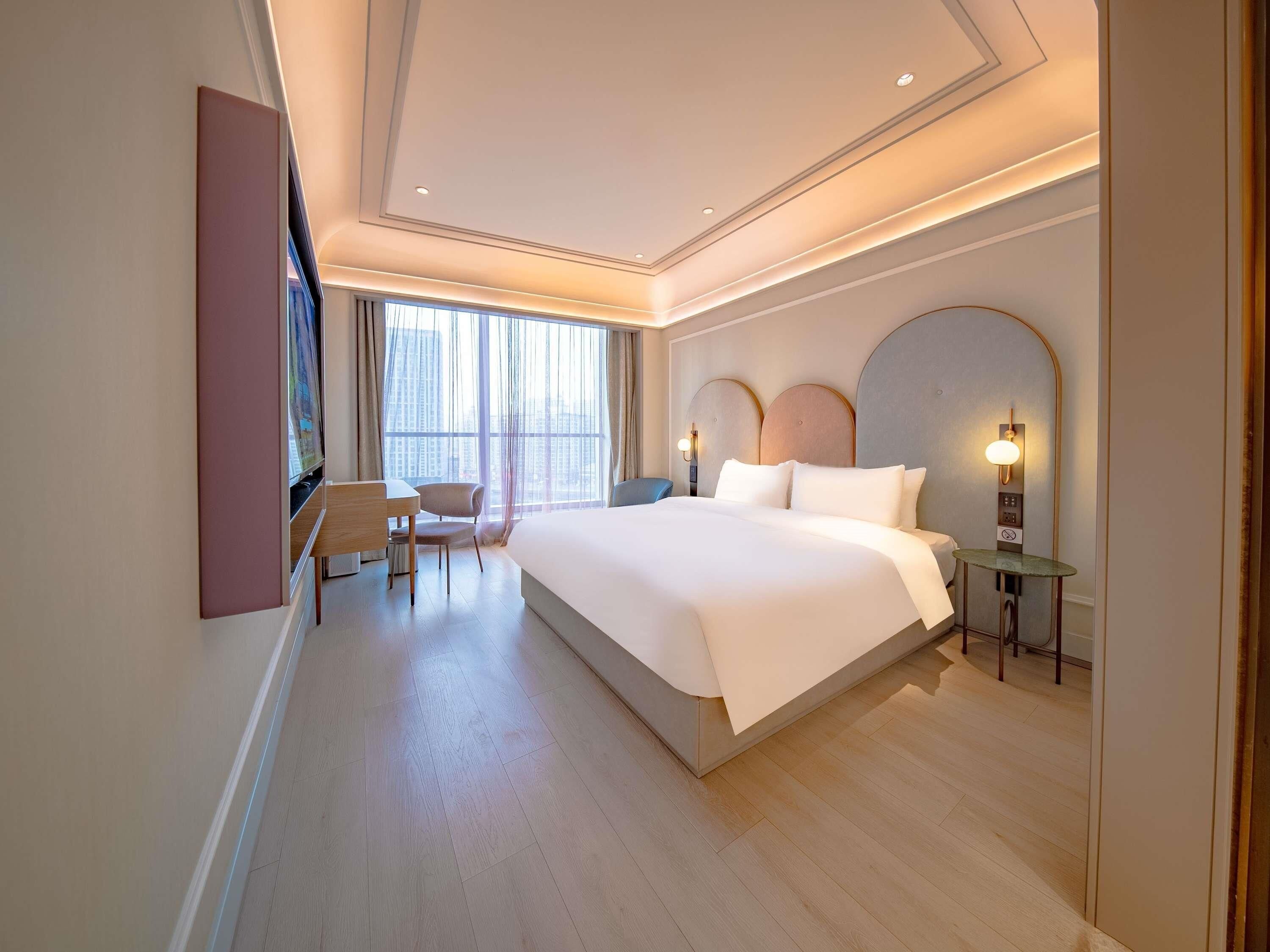 Habitación de hotel con una cama grande, un escritorio, una silla y vistas de la ciudad desde la ventana.