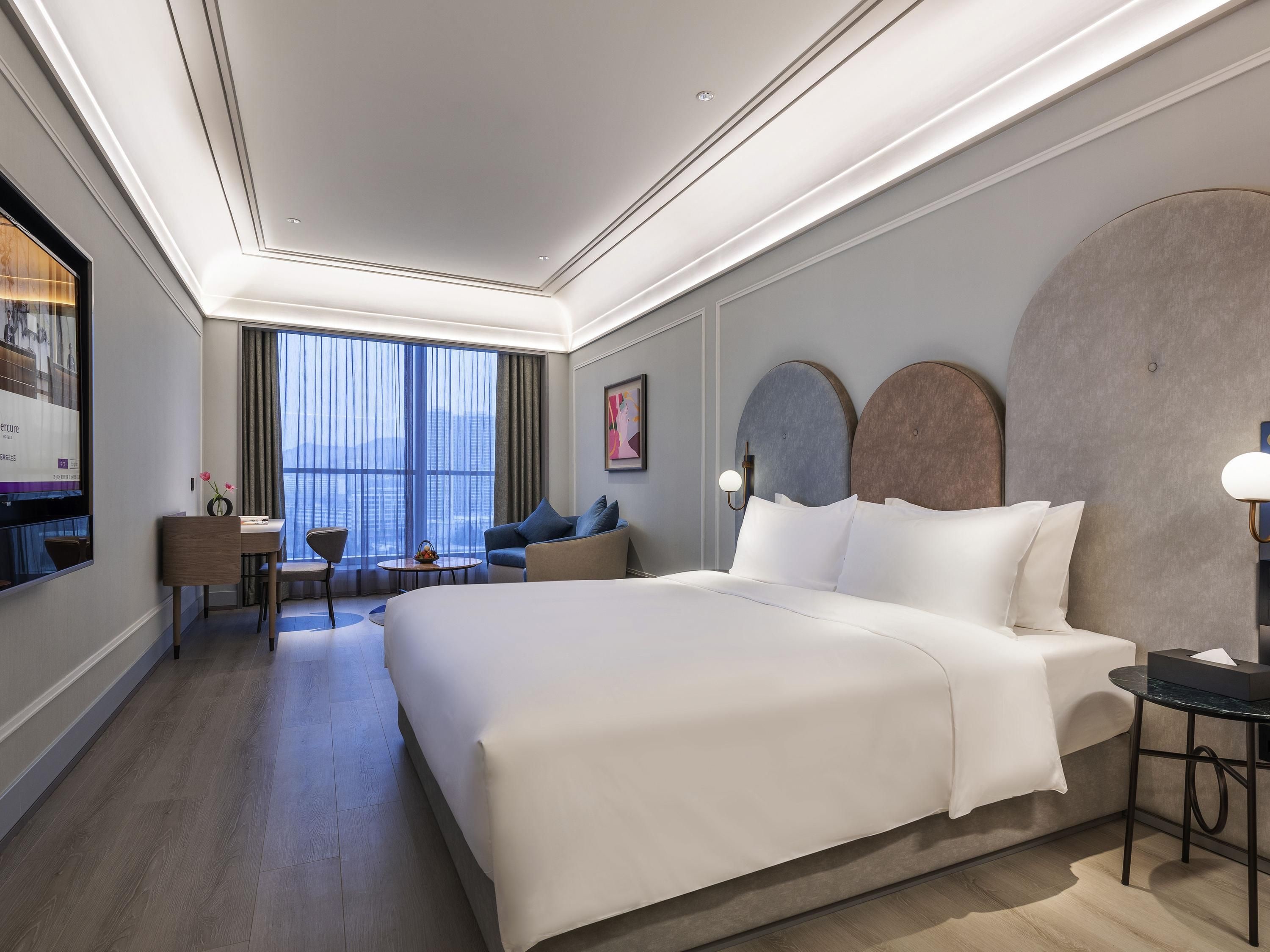Habitación de hotel moderna con una cama grande, un escritorio, una silla y vistas de la ciudad a través de amplios ventanales.