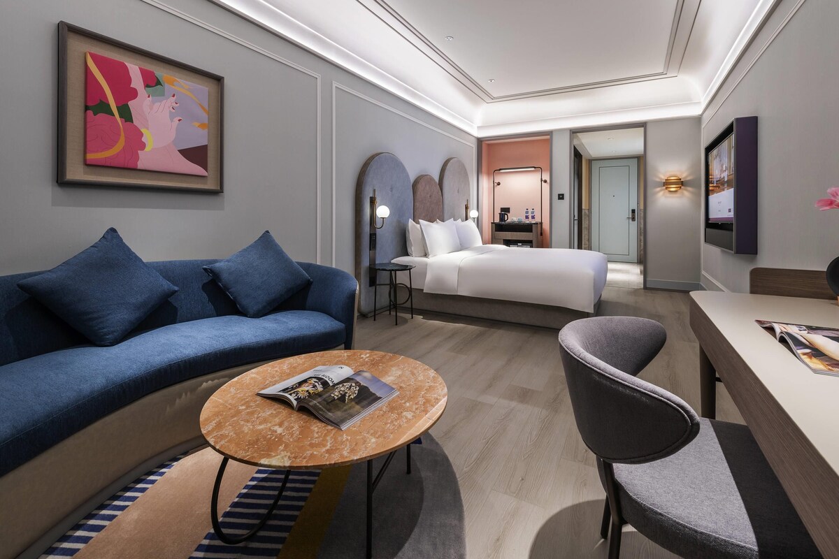 A modern hotel room with a blue sofa, a round marble table, a gray armchair, a bed, and a TV mounted on the wall.