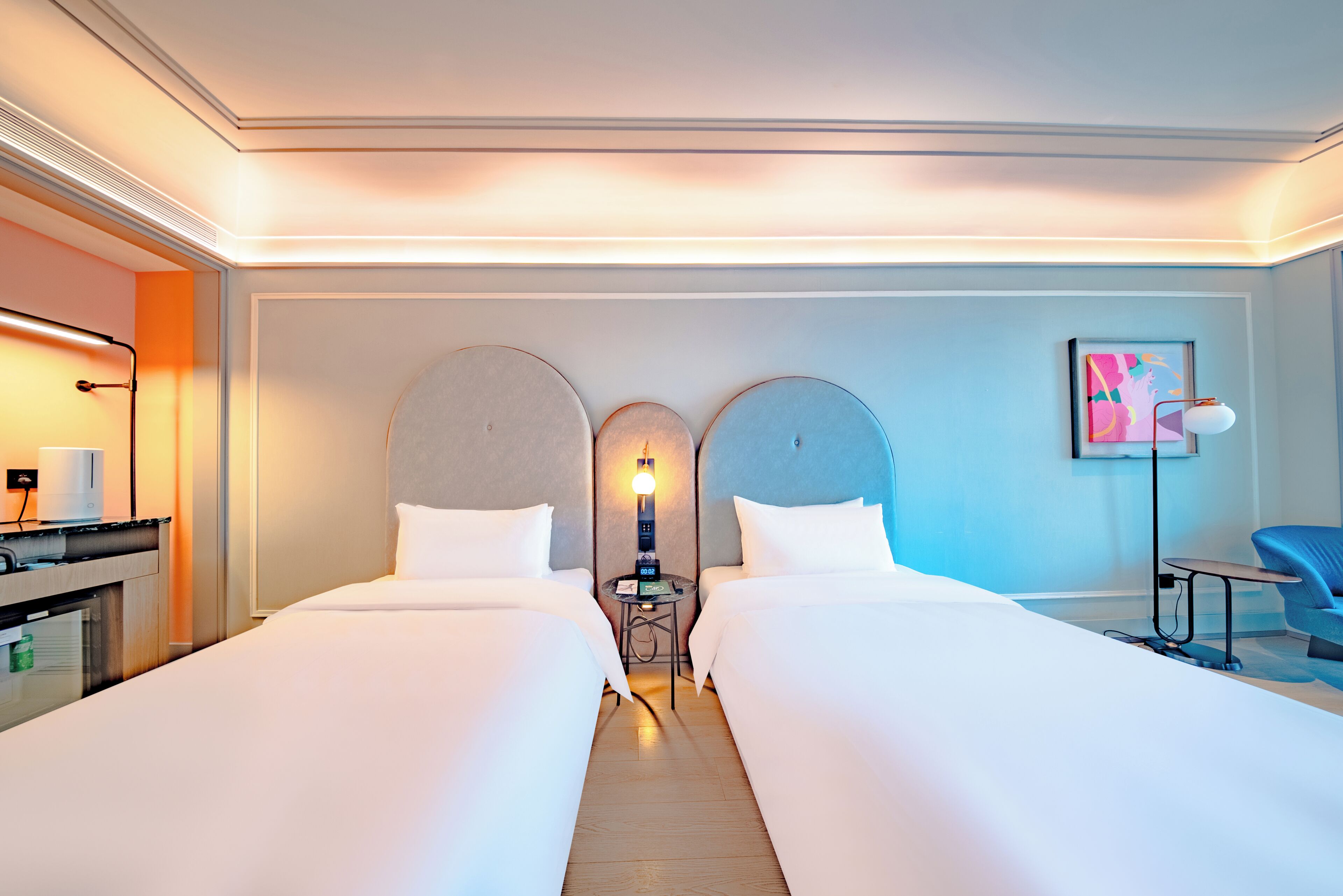 A hotel room with two beds, a desk, a chair, a lamp, and a painting on the wall.