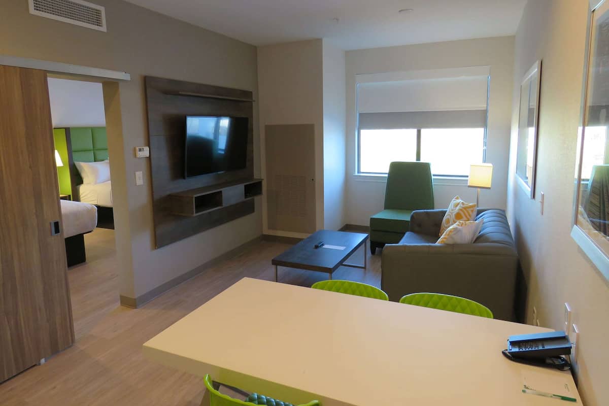 Een moderne hotelkamer met een flatscreen televisie aan de muur, een bank, een eettafel met stoelen en een raam met jaloezieën.