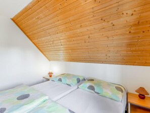 A room with a sloped wooden ceiling, a bed with patterned bedding, a bedside table with a lamp, and a wall-mounted shelf.