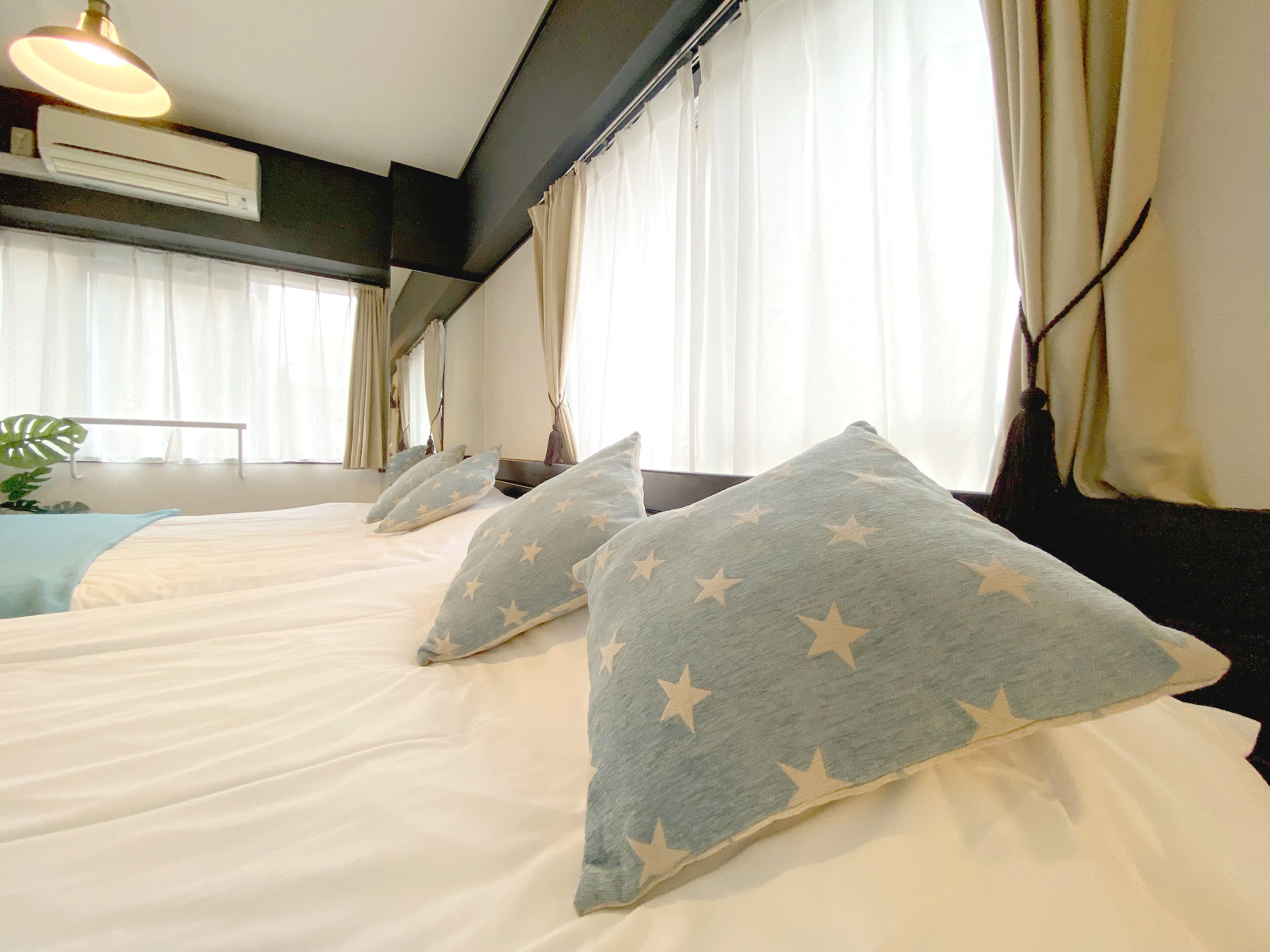 A hotel room with two beds, each with white bedding and star-patterned pillows. There is a ceiling fan, a potted plant, and a window with curtains.