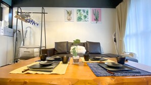 Una mesa de comedor dispuesta con platos, tazones y tazas, rodeada por un sofá y cojines, con una obra de arte en la pared y una ventana con cortinas.