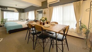 Una habitación de hotel con una cama, una mesa de comedor con sillas, una mesita con tazas y un florero, y un equipo de aire acondicionado.