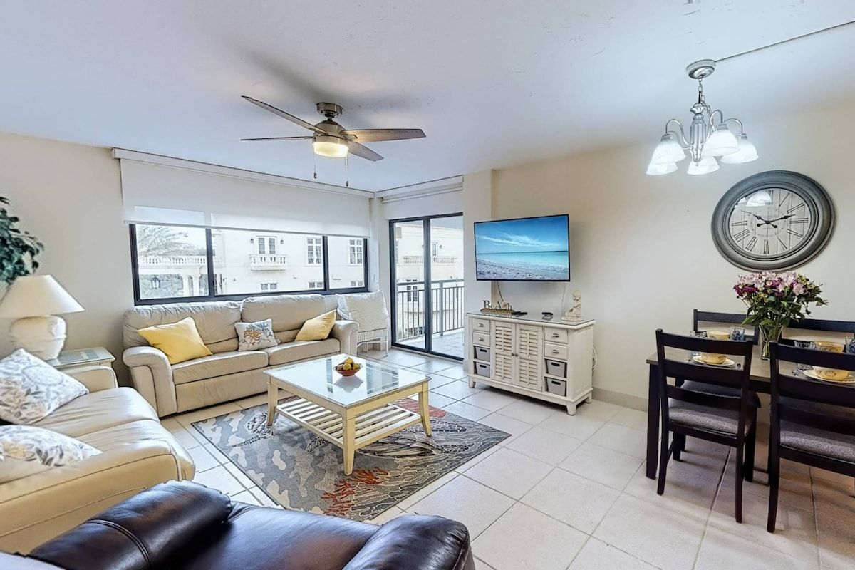 A living room with a sofa, coffee table, dining table with chairs, a ceiling fan, and a TV mounted on the wall.