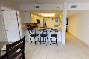 A kitchen with white cabinets, a microwave, and a refrigerator. There are three chairs at a small island with a sink. - Siesta Sands Private Beach | Gulf Views | Htd Pool (Sarasota)