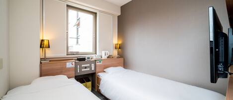 Habitación de hotel con dos camas, televisión, ventana con persianas y una mesita de noche con lámpara y taza.