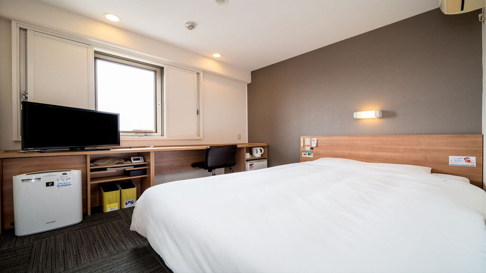 A hotel room with a bed, a desk with a TV, a small fridge, a chair, and a window.