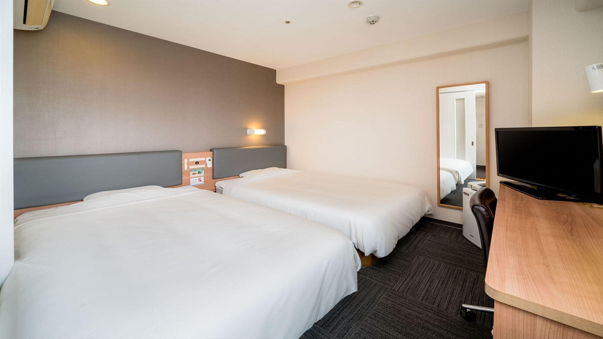 A hotel room with two beds, a desk with a TV, a mirror, and a wall-mounted phone.