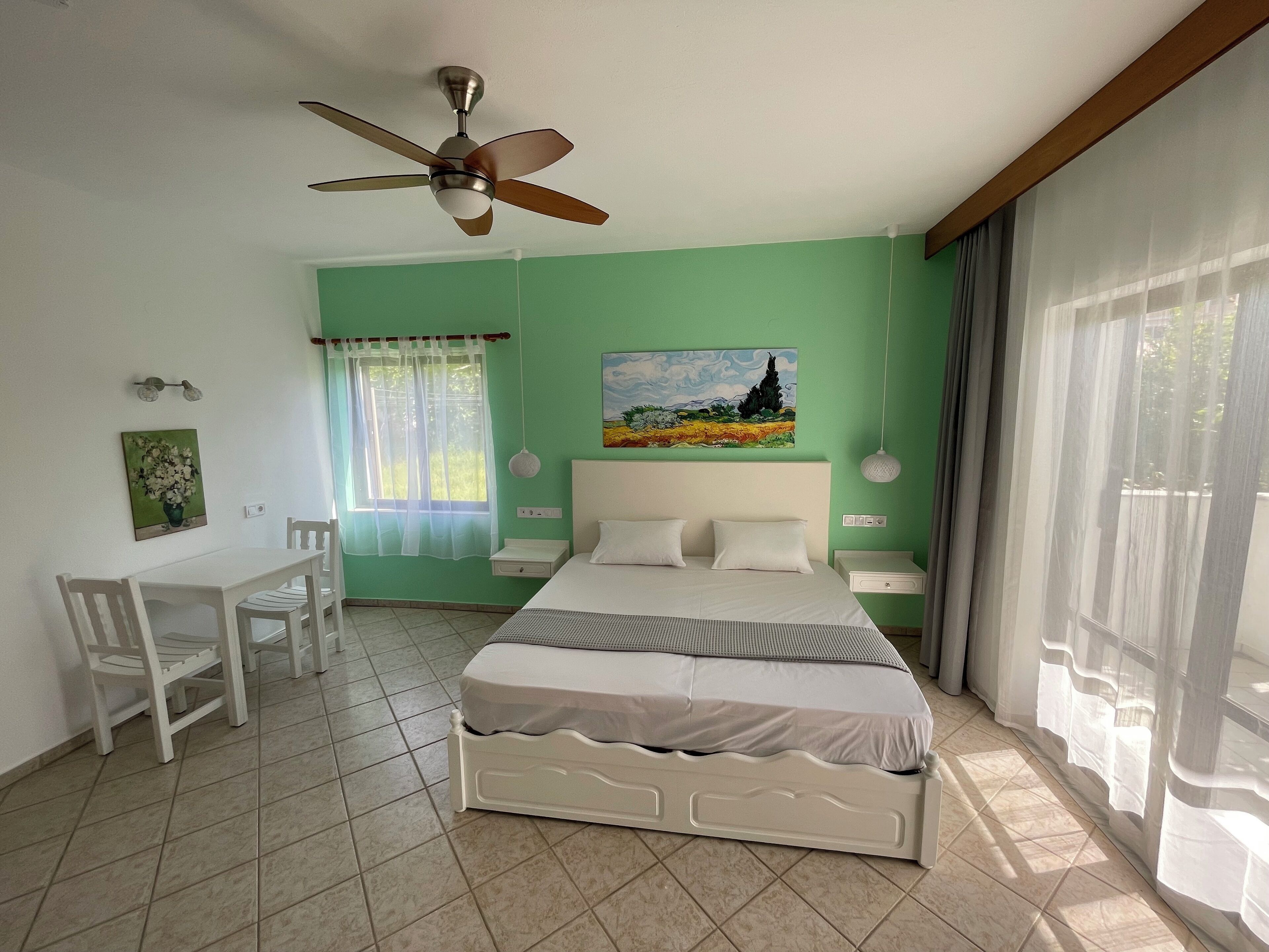 A bedroom with a bed, a desk, chairs, a ceiling fan, and a window with curtains.