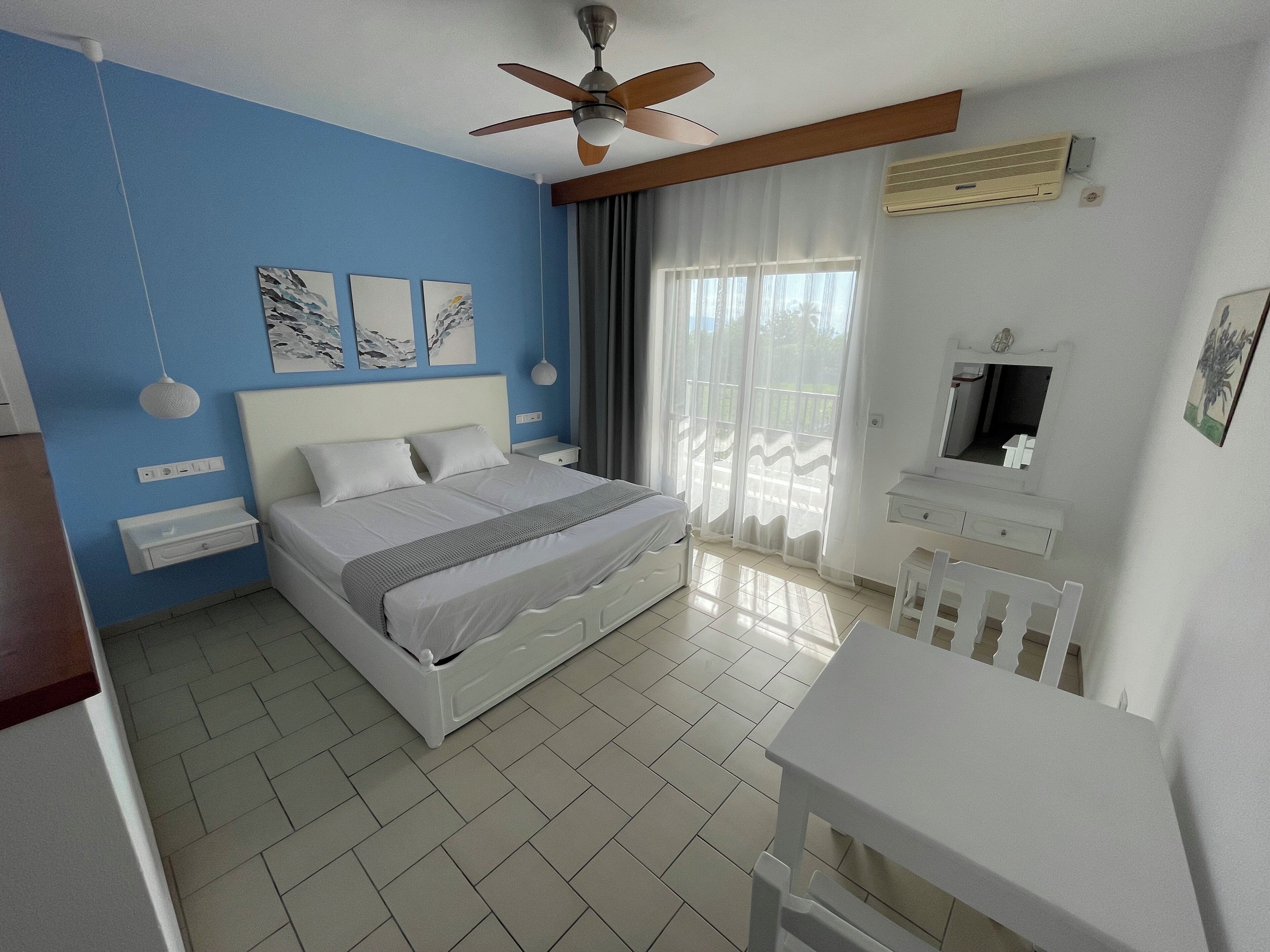 A bedroom with a bed, a desk, a chair, a ceiling fan, and a window with curtains.