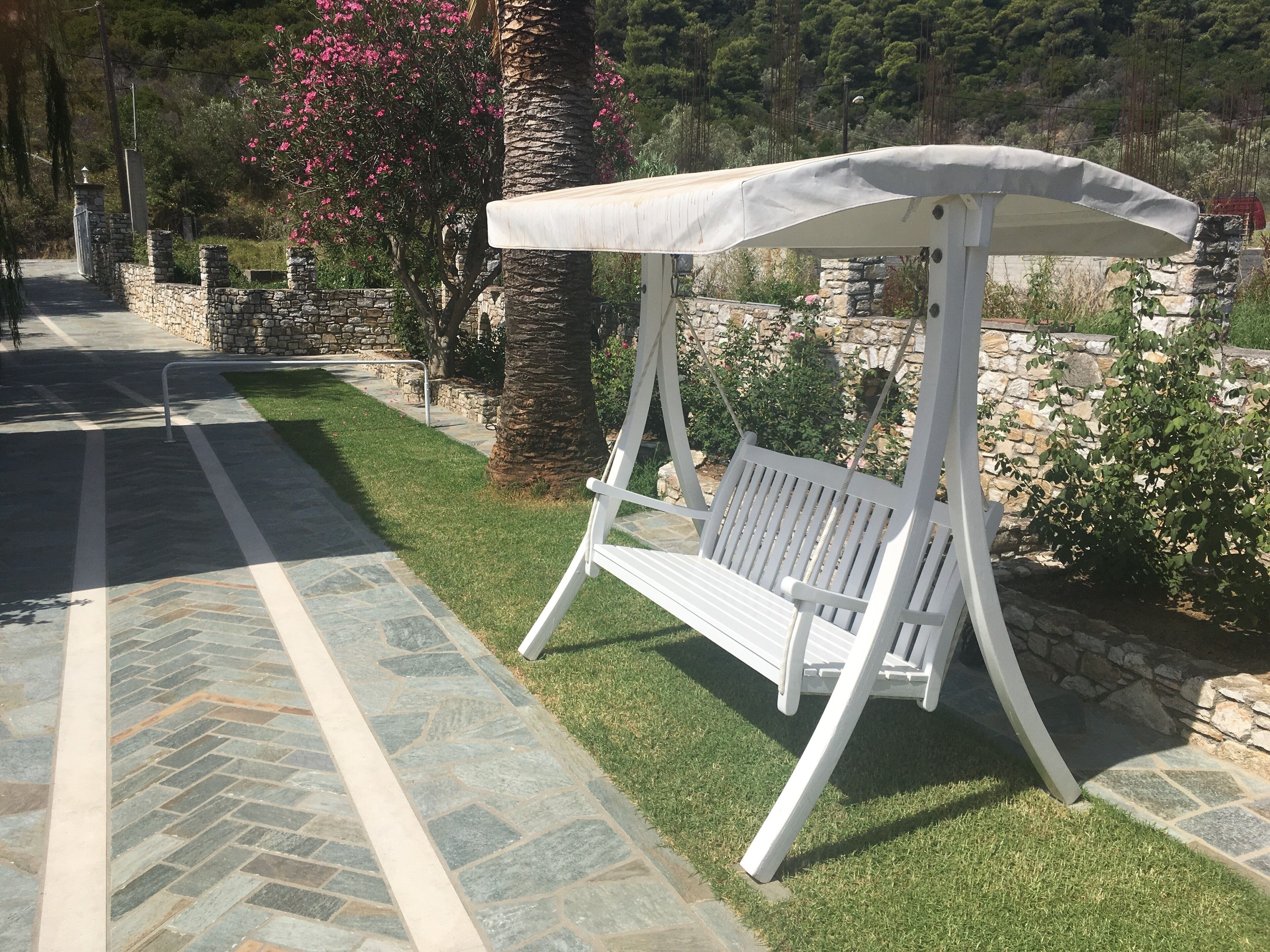 A white outdoor seating area with a bench and a canopy, surrounded by stone walls and greenery.