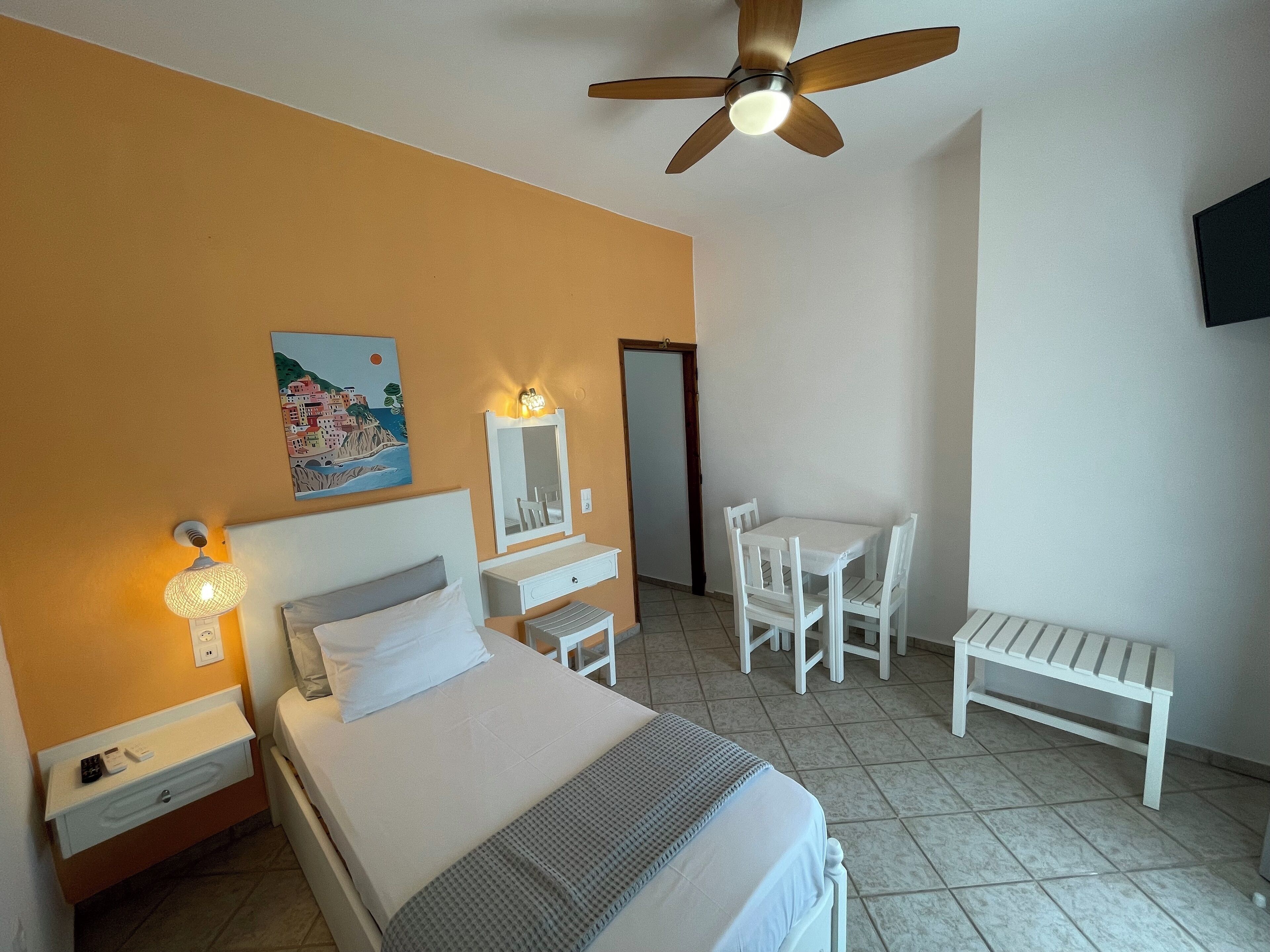 A hotel room with a bed, a desk, a chair, a ceiling fan, and a painting on the wall.