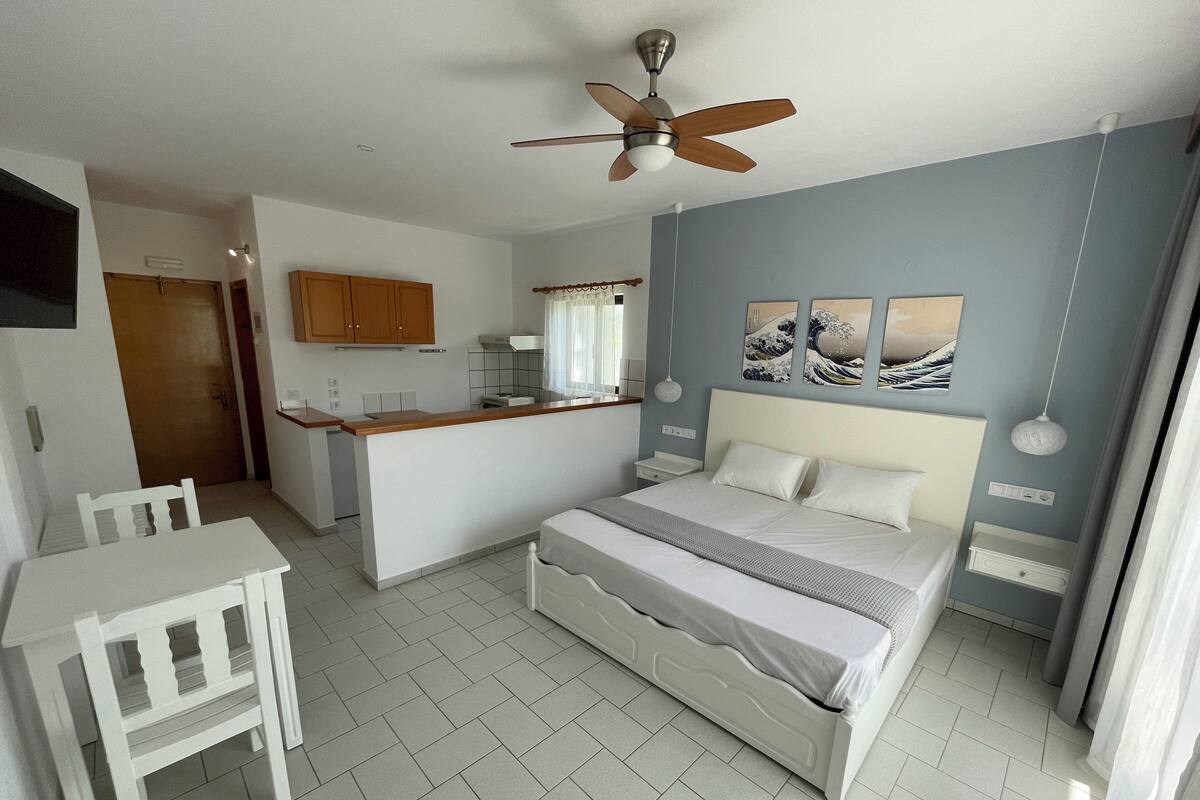 Quarto com cama, escrivaninha, cadeira, ventilador de teto e uma pequena cozinha compacta.