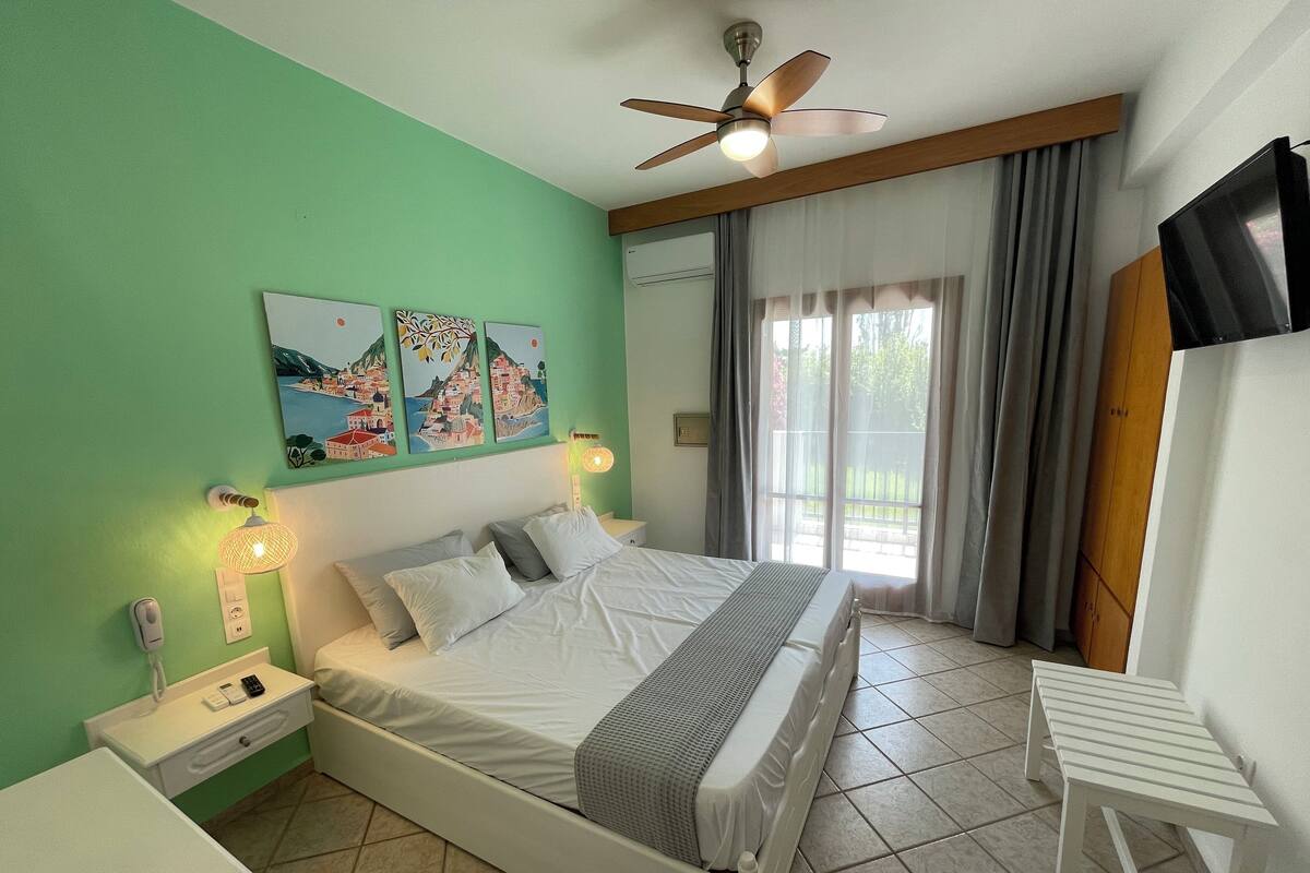 Quarto com cama, mesas de cabeceira, ventilador de teto, obra de arte na parede e vista pela janela.