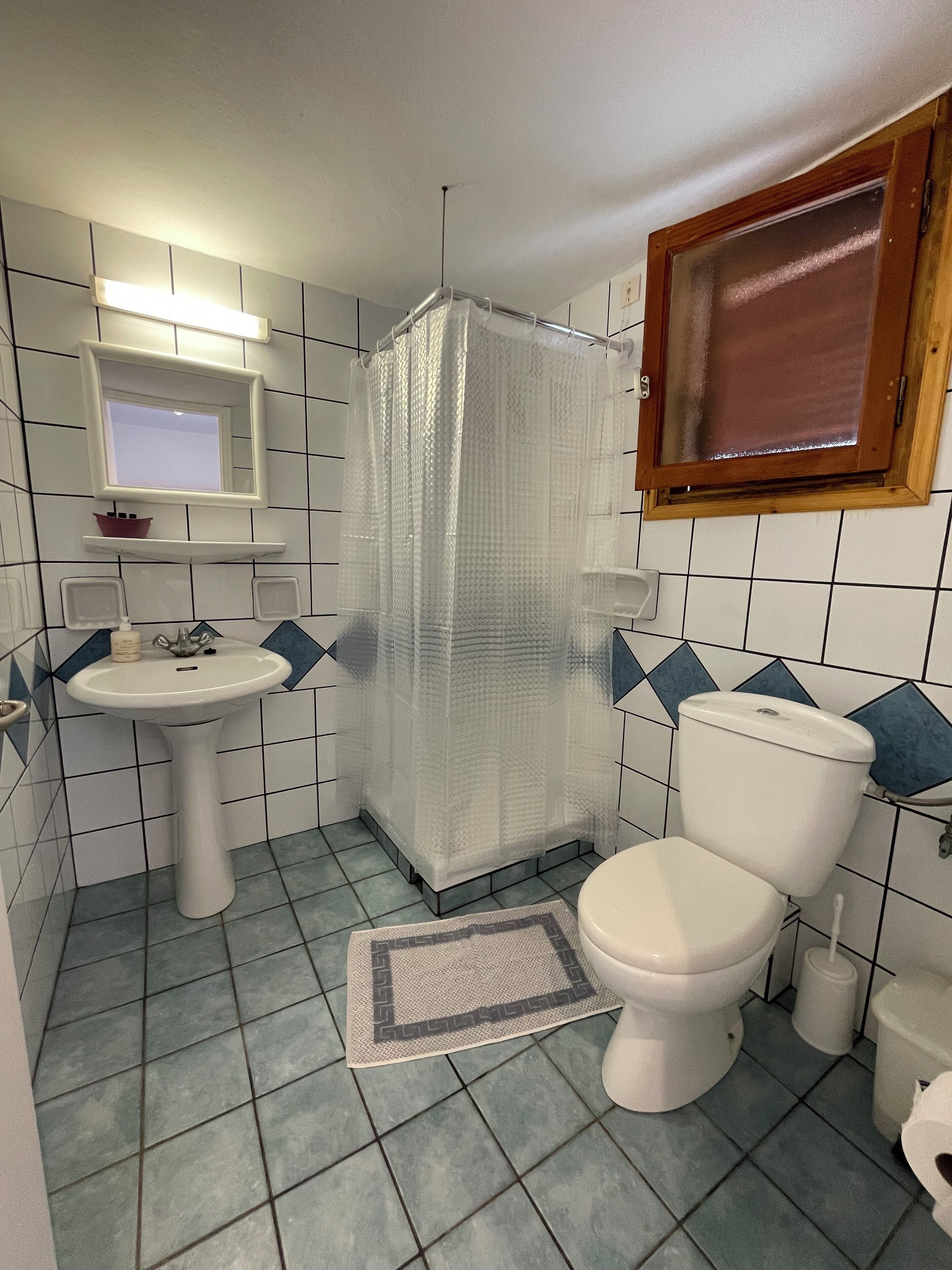 A bathroom with a toilet, sink, shower curtain, and a window above the toilet.