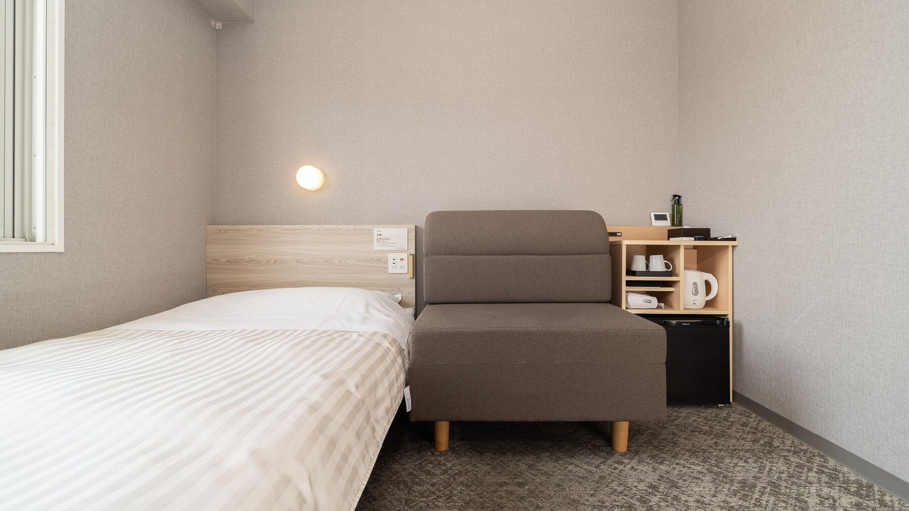 Una habitación de hotel pequeña y ordenada, con una cama individual, una silla, una mesita de noche con lámpara y un estante pequeño con una tetera y una taza.