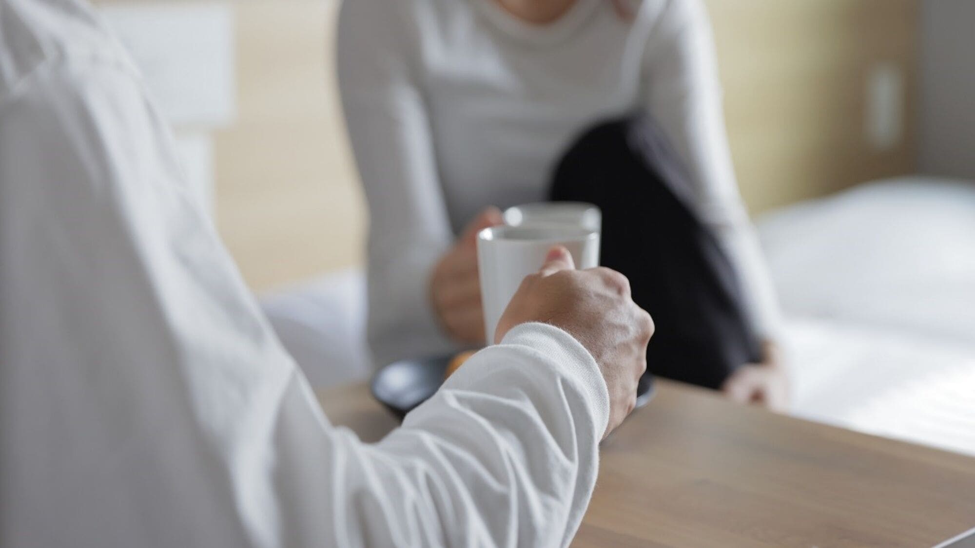 A person holding a glass of water at a table in a room with a bed and a nightstand.