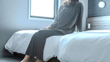 Eine Person sitzt auf einem Bett in einem Zimmer mit einem Fenster, einem Holz-Kopfteil und einem Lichtschalter an der Wand.