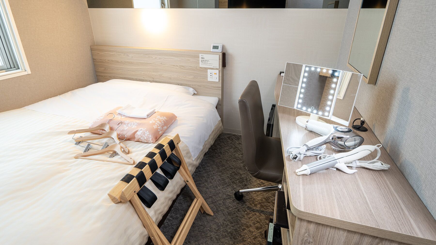 Habitación de hotel con una cama, una silla de madera, un escritorio con espejo e iluminación, y un reloj pequeño.