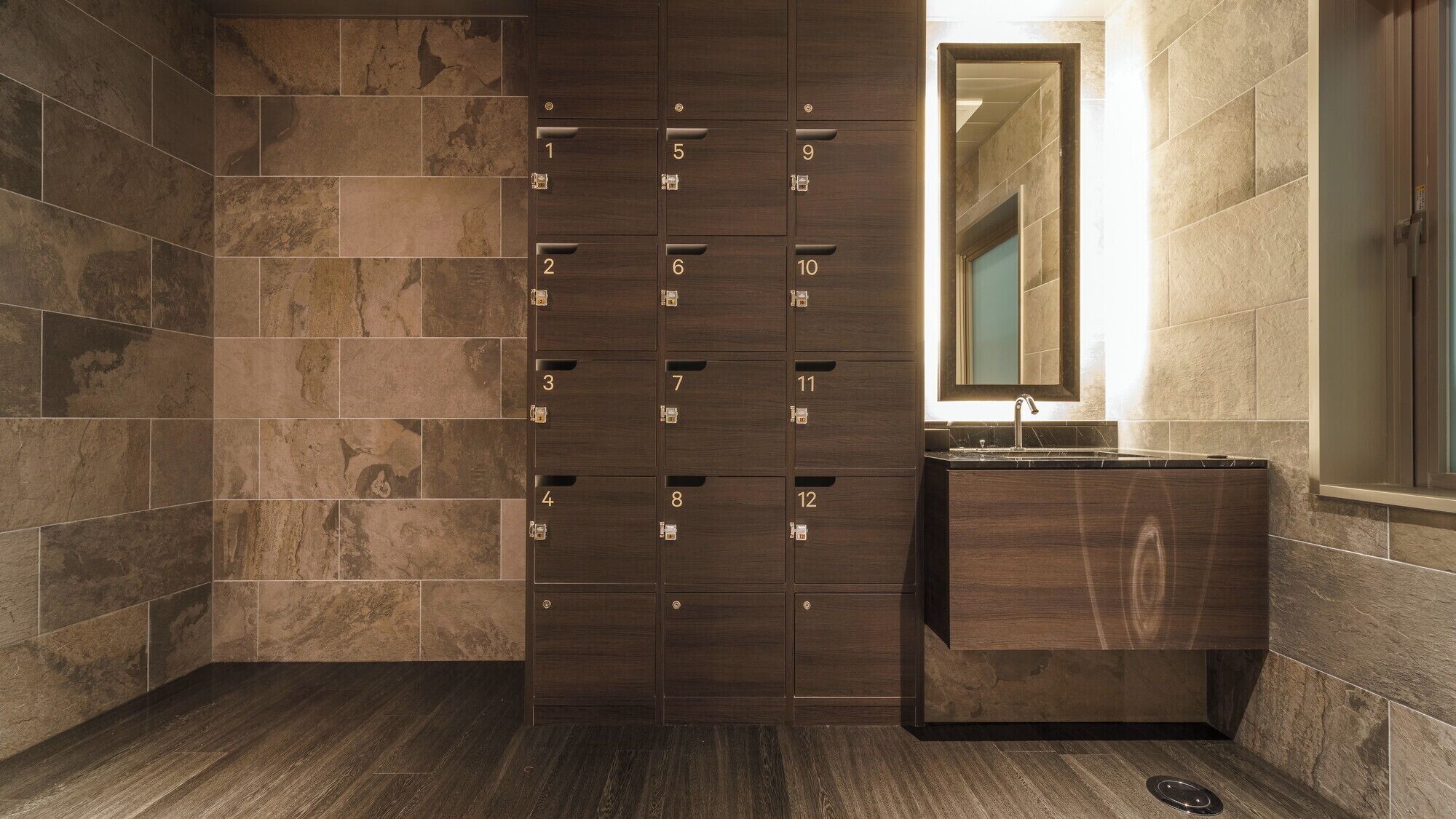 A modern bathroom with a dark wood vanity, a mirror above the sink, and a tiled wall with numbered lockers.