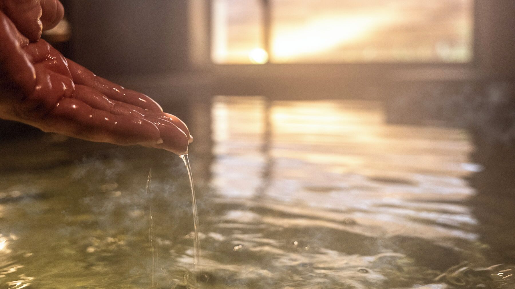 A hand holding a glass over a body of water with steam rising from it.