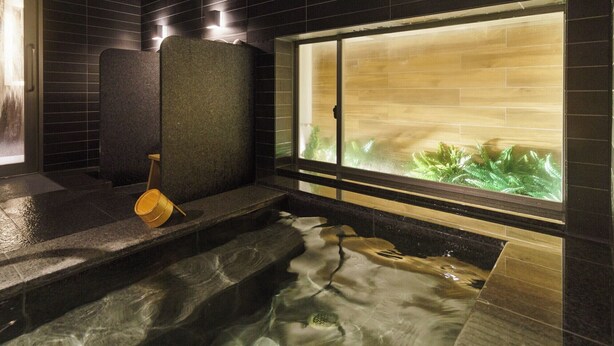 A hot spring bath with a wooden bucket, a stone bench, and a view of green plants through a window.