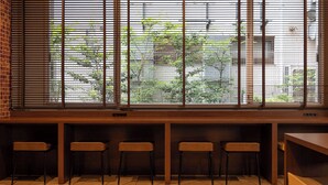 A seating area with four chairs and a wooden table, a window with metal blinds, and a view of greenery and buildings outside.
