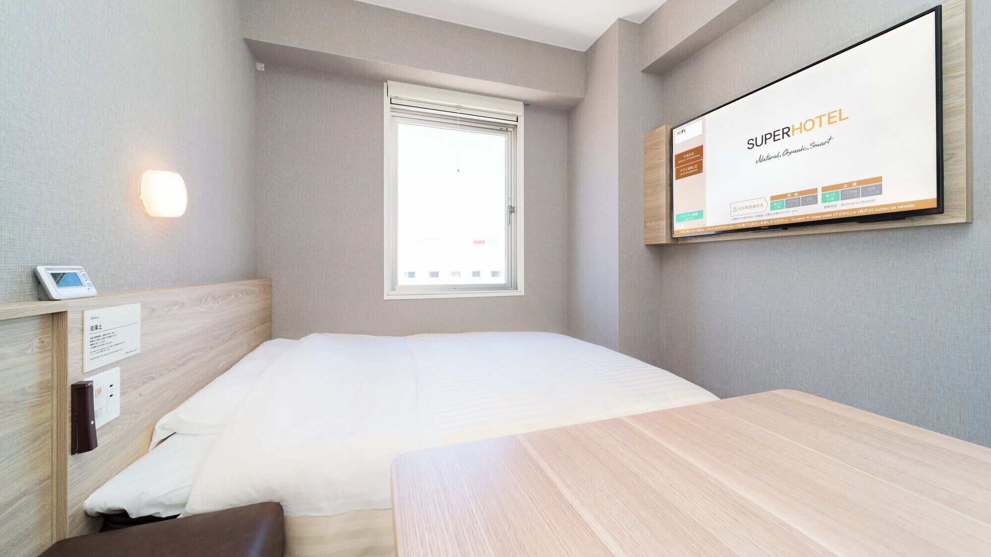 A hotel room with a bed, a desk, a TV mounted on the wall, and a window with blinds.