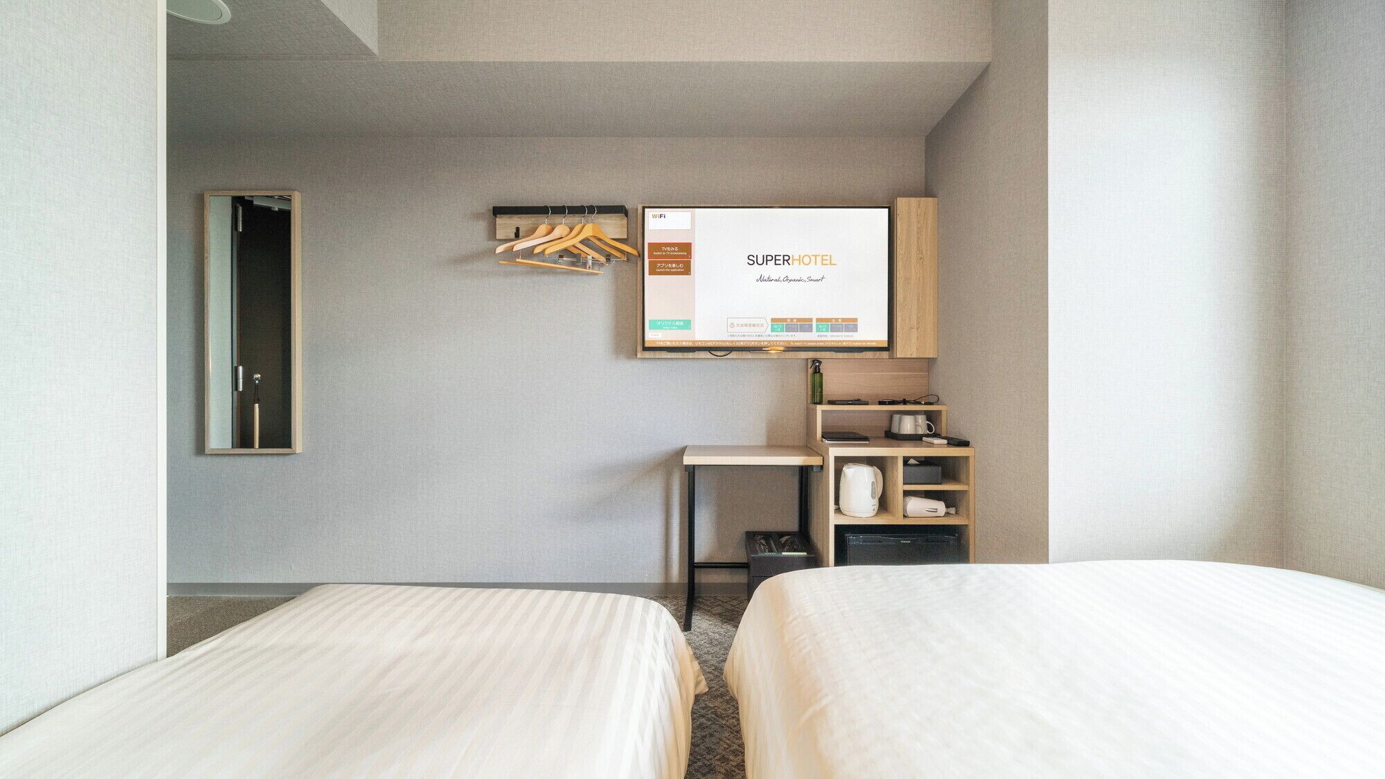 Una habitación de hotel con dos camas, un escritorio pequeño con un hervidor, un espejo y un televisor de pared mostrando 'SUPERHOTEL'.