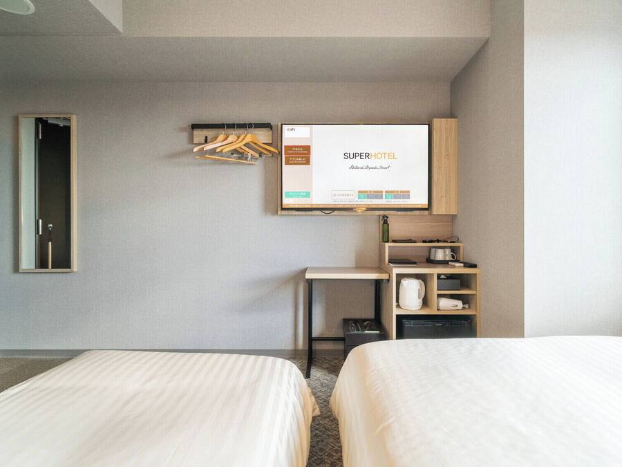 Una habitación de hotel con dos camas, un escritorio pequeño con un hervidor, un espejo y un televisor de pared mostrando 'SUPERHOTEL'.