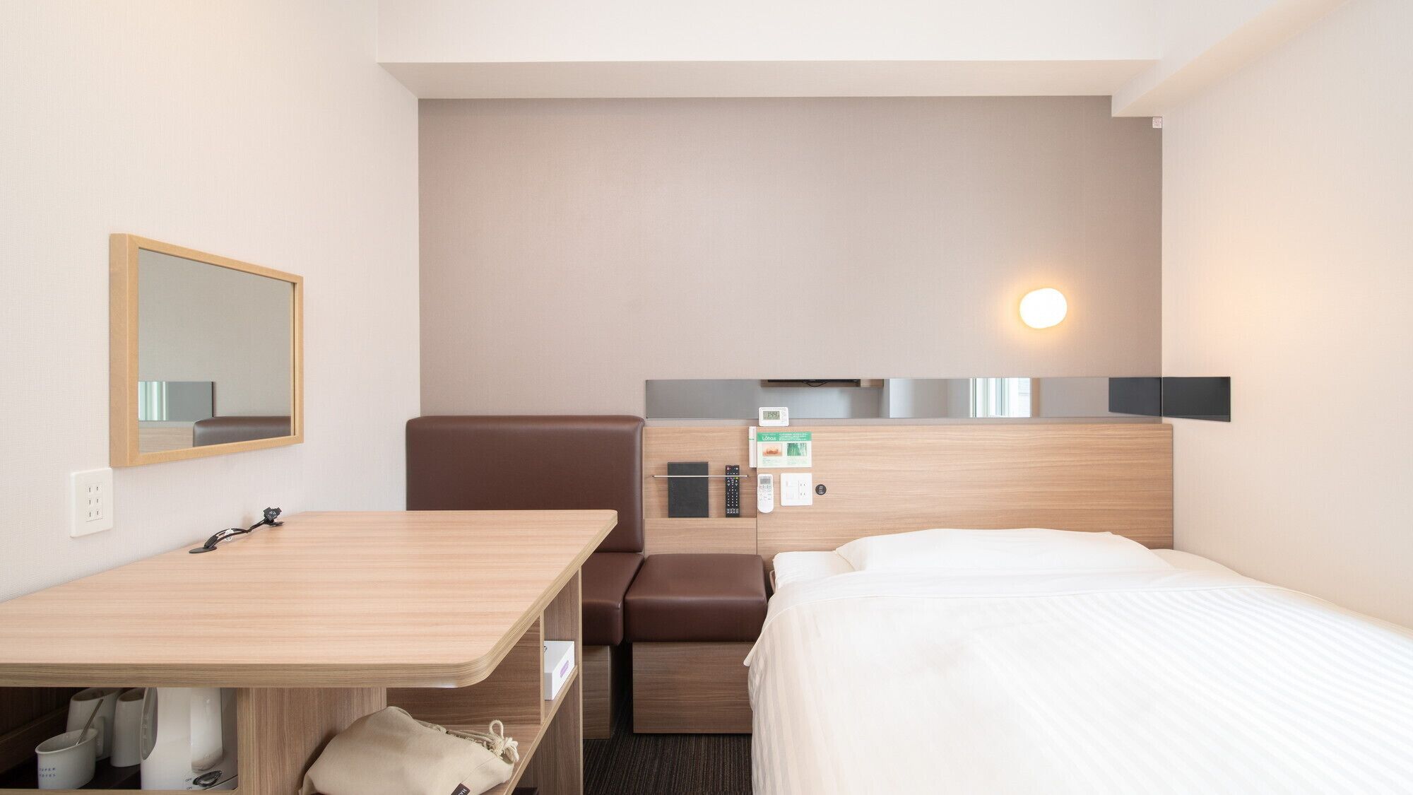 A hotel room with a bed, a desk, a chair, a mirror, a wall-mounted control panel, and a bedside table.