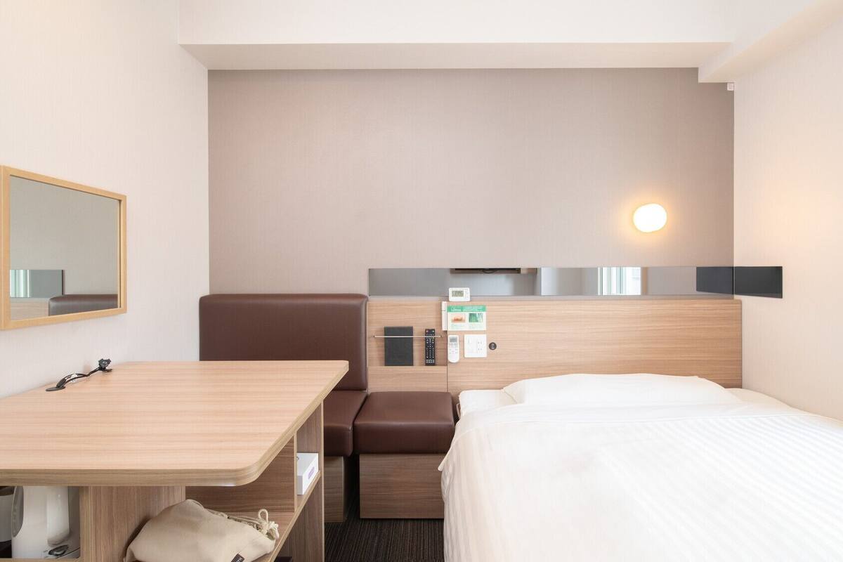 Habitación de hotel con cama, escritorio, silla, espejo, panel de control en la pared y mesita de noche.
