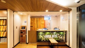 A hotel reception area with a wooden reception desk, a digital check-in kiosk, and a reception counter with a 'Super Hotel' sign. - Super Hotel Totsuka Station East (Yokohama)