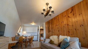 Una sala de estar acogedora con una pared de madera, un sofá con almohadones, un comedor con mesa y sillas, y una cocina al fondo.