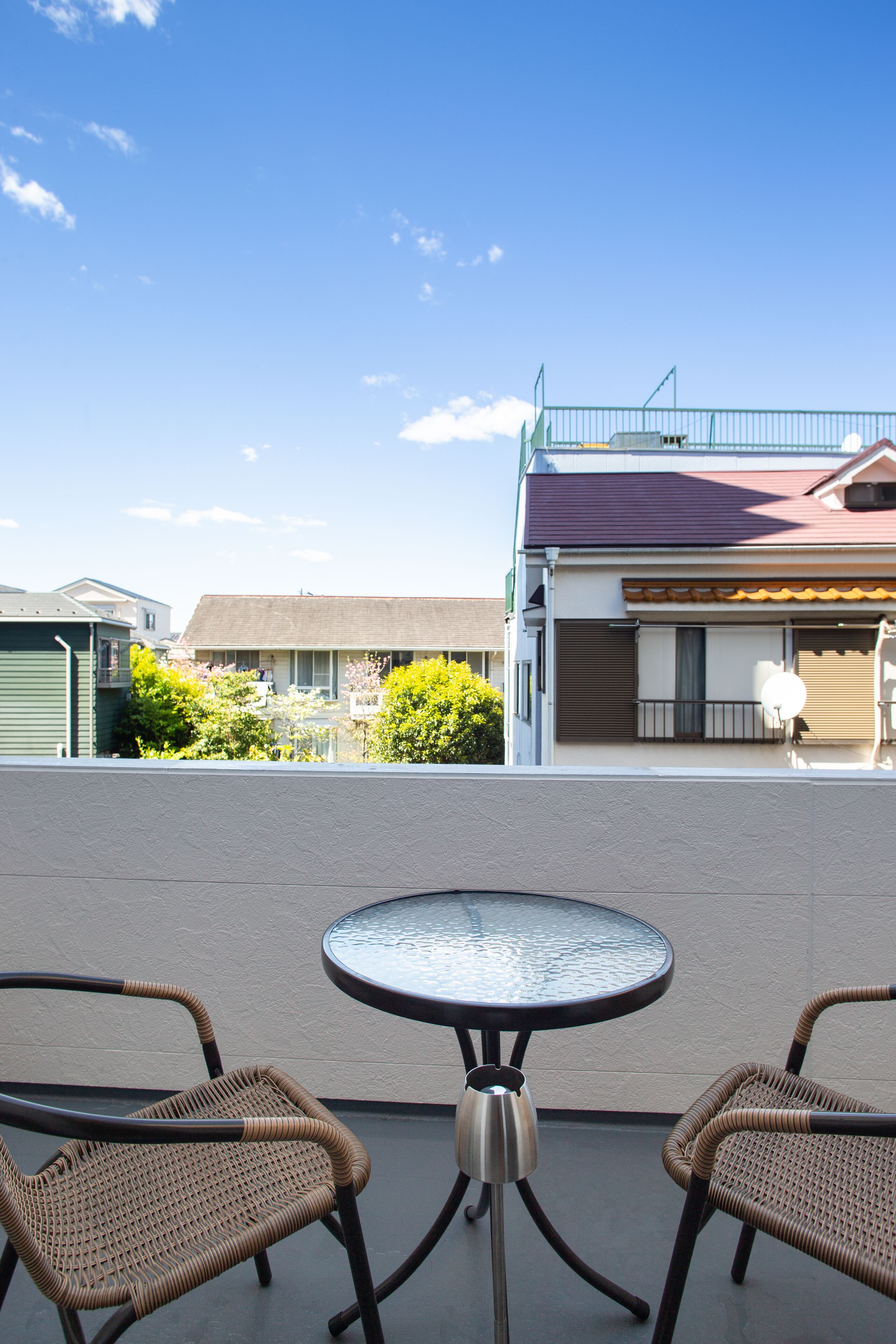 A balcony with two wicker chairs and a round table, overlooking a residential area with houses and greenery.