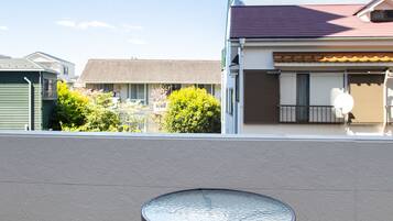 A balcony with two wicker chairs and a round table, overlooking a residential area with houses and greenery.
