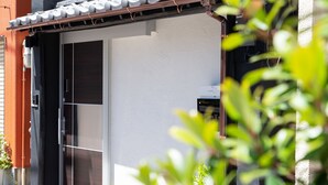 A building entrance with a sliding glass door, a small outdoor area with a potted plant, and a metal awning above the door. - My Home in Tokyo (Tokyo)